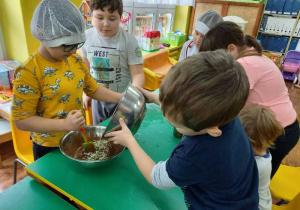 Przy stole dwóch chłopców miesza produkty mokre i sypkie. Jeden wsypuje suche produkty. Pozostałe dzieci przyglądaja się procesowi.