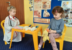 Chłopiec odgrywający rolę weterynarza siedzi na krzesełku i słucha objawów opisywanych przez dziewczynkę siedzącą również na krzesełku, która przyszła z kaczuszką do weterynarza. Pomiędzy dziećmi stoi stolik na którym znajdują się atrybuty weterynarza
