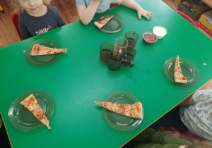 Dzieci siedzą przy stole. Na talerzykach leżą kawałki pizzy.