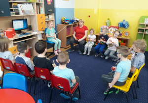 Dzieci siedzą na krzesełkach w kole przy biurku z komputerem. Trzymają w rękacg instrumenty.