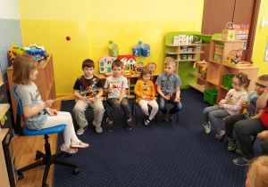Po lewej stronie na dużym krześle siedzi dziewczynka i trzyma batutę. Dyryguje grupą dzieci. Pozostałe dzieci siedzą na krzesełkach i grają na instrumentach.