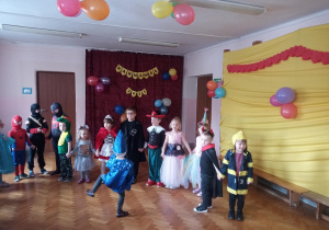 Widok na salę gimnastyczną. Prawa ściana ozdobiona żółtą tkaniną, na której zawieszone są balony. Na głównej ścianie napis "Bal karnawałowy". Na środku sali dzieci w przebraniach tańczą.