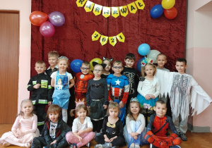 Dzieci z grupy 1 w przebraniach karnawałowych. Za nimi na ścianie napis "Bal karnawałowy".
