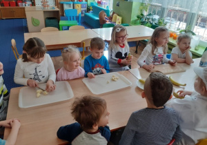 Dzieci siedzą przy stołach. Jedna dziewczynka kroi banany. Chłopiec ma przed sobą tacę z pokrojonymi już bananami.