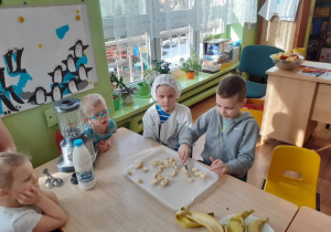 Czworo dzieci siedzi przy stole. Chłopiec kroi banany, reszta dzieci obserwuje.