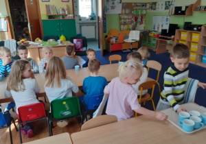 Dzieci siedzą przy stole. Dwoje pełniących rolę kelnerów odkłada na tacę kubki po wypitym koktajlu.