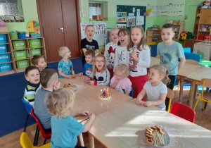 Dziewięcioro dzieci siedzi przy stołach, pięcioro stoi przy stole. Na stole znajduję się tort i talerze ze słodyczami.