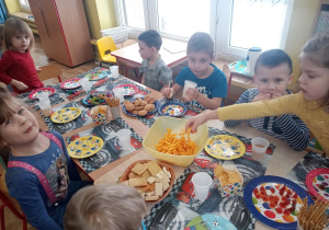 Przy złączonych stołach, na których ustawione są talerze z przysmakami, siedzą dzieci.