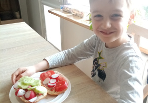 Po prawej stronie, przy stole siedzi chłopiec. Na stole leży talerz. Na talerzu kanapki z warzywami.