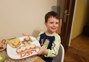 Po prawej stronie, przy stole siedzi chłopiec. Na stole leży talerz. Na talerzu kanapki z warzywami.