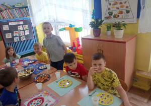 chłopiec stoi przy stoliku, pozostałe dzieci siedzą przy stolikach, świętują urodziny kolegi