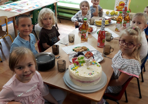 widok na stoły, przy których siedzą stoły, na stole tort, talerze, kubki, słodycze, napoje