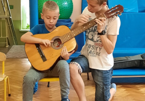 Chłopiec siedzi na krzesełku i próbuje grać na gitarze z pomocą starszego kolegi.