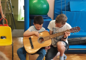 Chłopiec siedzi na krzesełku i próbuje grać na gitarze z pomocą starszego kolegi.