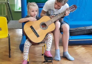 Dziewczynka siedzi na krzesełku i próbuje grać na gitarze z pomocą starszego kolegi.