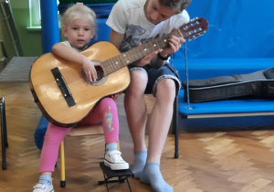 Dziewczynka siedzi na krzesełku i próbuje grać na gitarze z pomocą starszego kolegi.