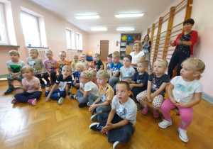 Dzieci siedzą na podłodze i na ławeczkach słuchając koncertu.