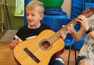 Chłopiec siedzi na krzesełku i próbuje grać na gitarze z pomocą starszego kolegi.