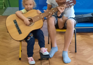 Dziewczynka siedzi na krzesełku i próbuje grać na gitarze z pomocą starszego kolegi.