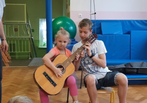 Dziewczynka siedzi na krzesełku i próbuje grać na gitarze z pomocą starszego kolegi.