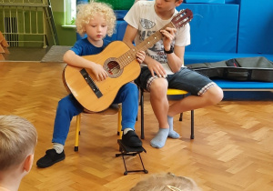 Chłopiec siedzi na krzesełku i próbuje grać na gitarze z pomocą starszego kolegi.