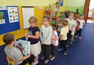 Chłopiec siedzi na krześle, przed nim dzieci ustawione w kolejce składają mu życzenia.