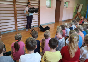 Przedszkolaki siedzą na sali gimnastycznej i słuchają koncertu w wykonaniu uczennicy szkoły muzycznej, która gra na waltorni -rogu.