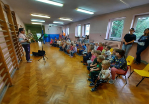Przedszkolaki siedzą na sali gimnastycznej i słuchają koncertu w wykonaniu uczennicy szkoły muzycznej, która gra na waltorni -rogu.