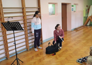 Pani od rytmiki prezentuje torbę w której znajduje się waltornia. Za nią stoi dziewczynka.