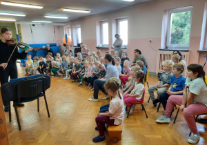 Dzieci siedzą i słuchają koncertu w wykonaniu uczennicy, która gra na skrzypcach.