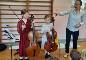 Uczennice stoją i prezentują swoje instrument. Pani od rytmiki wyjaśnia dzieciom jak zbudowana jest wolonczela.