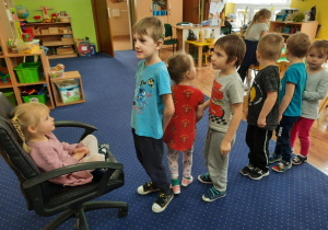 Dziewczynka siedzi na krześle, a dzieci ustawione w kolejce i składają jej życzenia.