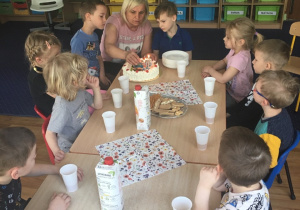 Dzieci siedzą przy stole, a pani wkłada świeczki w tort.