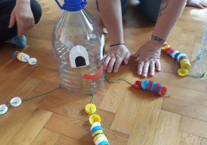 Na podłodze stoi plastikowa butelka ze sznurkami na których są nawleczone korki.