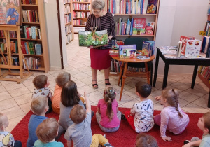 dzieci siedzą na czerwonym dywanie w bibliotece, pani bibliotekarka pokazuje książki