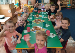 Dzieci siedzą przy stole i machają ręką.