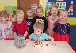 Chłopiec siedzi przy stole przed tortem i zdmuchuje świeczki, a pozostałe dzieci stoją za nim.