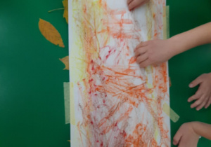 Dzieci kolorują karton kredkami pastelowymi pod którym leżą liście.