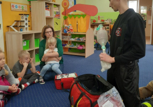 Dzieci siedzą na dywanie i oglądają wyposażenie torby strażaka.