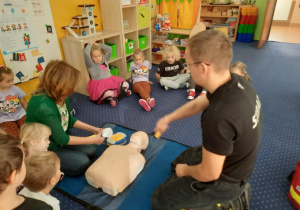 Dzieci siedzą na dywanie i uczą się jak należy udzielać pierwszej pomocy przedmedycznej.