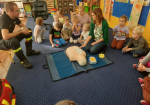 Dzieci siedzą na dywanie i uczą się jak należy udzielać pierwszej pomocy przedmedycznej.
