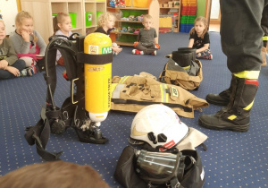 Dzieci siedzą na dywanie i oglądają wyposażenie stroju strażaka.