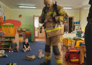Dzieci siedzą na dywanie i obserwują jak strażak ubiera się w swój codzienny strój do akcji.