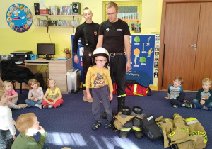 strażacy pokazują dzieciom sprzęt do ratowania życia i gaszenia pożarów, dzieci przymierzają hełm strażacki