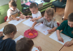 Dzieci siedzą przy stole malują palcem umoczonym w farbie literę "I, i".