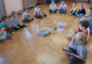 Dzieci siedzą w półkolu i na różnych instrumentach grają jesienną muzykę.