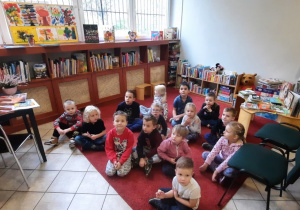 Dzieci siedzą na dywanie w bibliotece i słuchają pani.