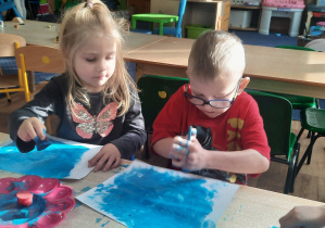Dzieci siedzą przy stole i stemplują karton niebieską farbą.