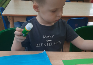 Chłopiec siedzi przy stole i nakleja zielony papier na karton.