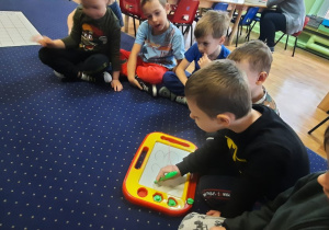 Dzieci siedzą na dywanie w kole, chłopiec pisze cyfrę 3.